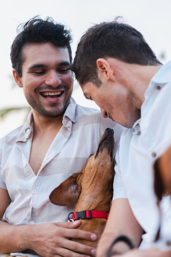 two men standing together as a couple, one is holding a small brown dog that is looking up at the other man